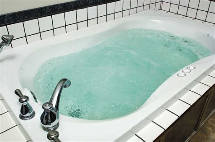 A full bathtub topped with a bath tray holding a glass of wine, a tablet, a phone, and a mug of coffee